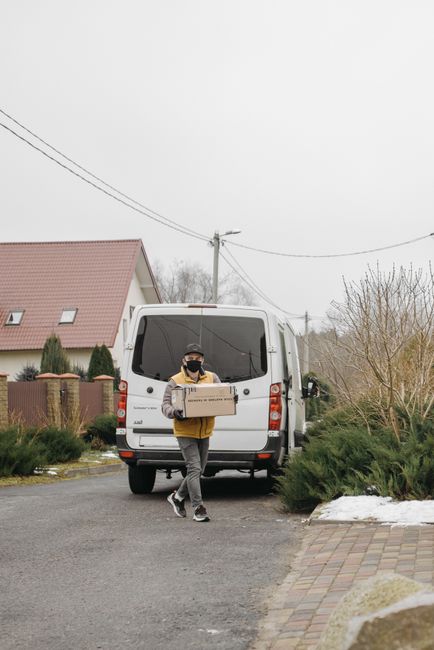 A young male mover stands confidently with arms crossed in front of a white delivery van branded with the words 'DELIVERY ALWAYS ON TIME,' parked on a residential street. The van is positioned beside
