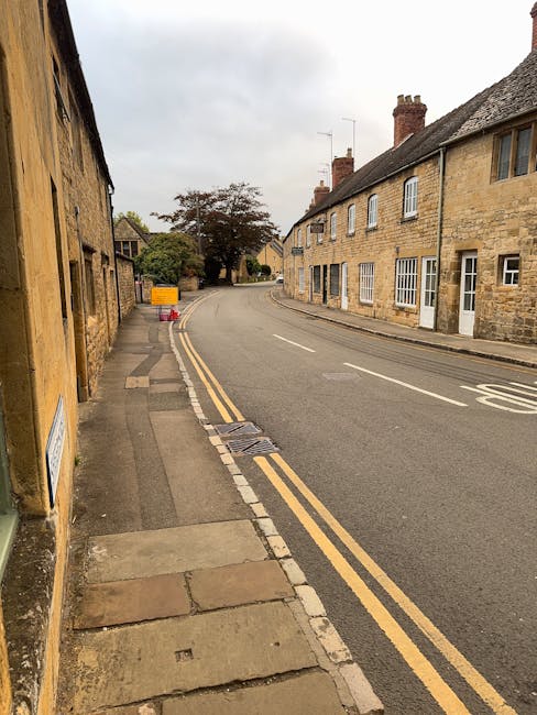 An empty residential street with a gentle curve, lined by traditional stone-built houses with slate roofs, some featuring brick chimneys. The pavement on the left side includes a narrow sidewalk with 