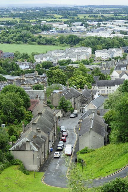 A wide aerial view of a residential area with rows of closely packed houses featuring grey rooftops and stone facades, surrounded by lush green trees and grassy areas. Narrow streets with parked cars