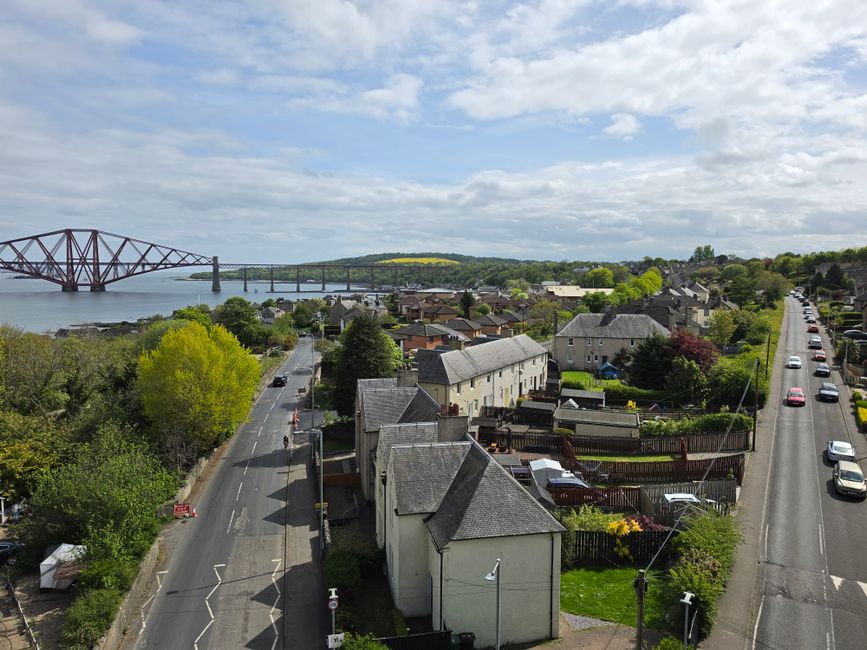 A wide aerial view of a residential area with rows of closely packed houses featuring grey rooftops and stone facades, surrounded by lush green trees and grassy areas. Narrow streets with parked cars