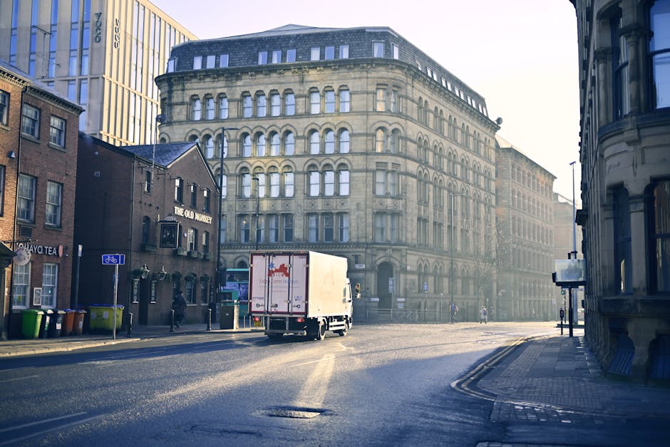 A white box truck with a red logo is parked on a city street near a kerb, ready for home relocation or furniture transport. The truck is adjacent to older and modern buildings, with one prominent multi-storey stone building featuring numerous windows and decorative arches. Next to the truck, on the pavement, there are several large cardboard boxes wrapped in plastic, likely packed for a house move, and a couple of moving blankets or protective coverings. The scene shows a quiet urban environment during daylight with some pedestrians and cyclists in the background. The street has a bicycle lane marked with a blue sign, and nearby, a row of colored waste bins is visible. The surroundings include shopfronts, street signage, and streetlights, all contributing to the typical urban setting for a professional removals service, such as Man and Van Queensbury, involved in packing and moving activities near residential and commercial properties.
