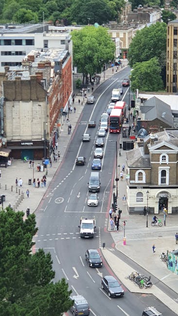 An urban street scene viewed from an elevated perspective, showing a row of parked cars along the curb and moving vehicles on a two-lane road, with pedestrians walking on the sidewalk and crossing at designated crossing points. The street is lined with a mix of commercial and residential buildings, including a brick building with signage and a smaller white structure with arched windows. Green trees are scattered along the street, providing shade and greenery amidst the buildings. The scene appears to be during daytime with bright natural light, and the overall environment depicts a busy, well-developed area suitable for home relocation or furniture transport logistics, as part of a professional removal service by Man and Van Queensbury. The image captures the typical setting for loading and unloading furniture during a house move, demonstrating the transportation of household goods in an active urban neighbourhood.